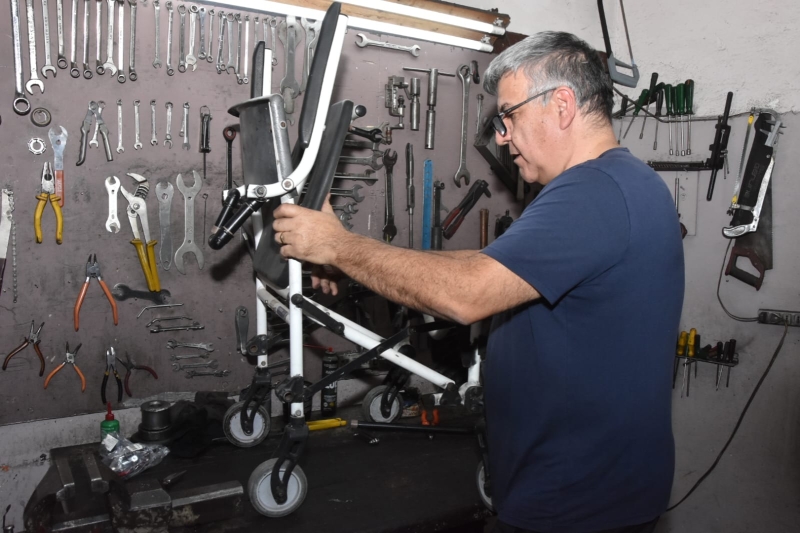 Descrição: A imagem mostra uma pessoa em uma oficina de manutenção ajustando uma cadeira de rodas dobrável sobre uma bancada de trabalho. A pessoa está em pé, de perfil, usando camiseta azul-marinho e óculos, com as duas mãos segurando a cadeira enquanto parece verificar ou realizar um reparo. Ao fundo, há uma grande parede organizada com diversas ferramentas penduradas, como chaves de boca, alicates, chaves de fenda, serras e martelos, indicando um ambiente profissional voltado para consertos mecânicos. Sobre a bancada também há peças, ferramentas e pequenos componentes espalhados, reforçando a ideia de que a cadeira está passando por manutenção. O espaço é simples, bem iluminado e transmite a impressão de um local dedicado ao reparo de equipamentos de mobilidade. Fim da descrição.