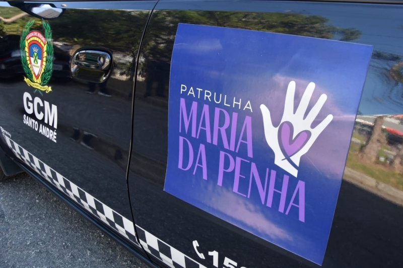 descrição: A imagem mostra a lateral de uma viatura preta da Guarda Civil Municipal (GCM) de Santo André, fotografada de perto. À esquerda, está o brasão da corporação, acompanhado da inscrição "GCM Santo André" em letras brancas. No centro da porta, há um grande adesivo retangular em tons de azul e roxo com a frase "Patrulha Maria da Penha". Ao lado do texto, aparece o desenho de uma mão branca aberta, com um coração roxo na palma, símbolo da campanha de proteção às mulheres em situação de violência doméstica. Na parte inferior da viatura há uma faixa xadrez em preto e branco, característica de veículos oficiais. O reflexo de árvores, da rua e de carros ao redor pode ser visto na pintura brilhante do veículo, indicando que a foto foi feita ao ar livre, durante o dia. Fim da descrição.
